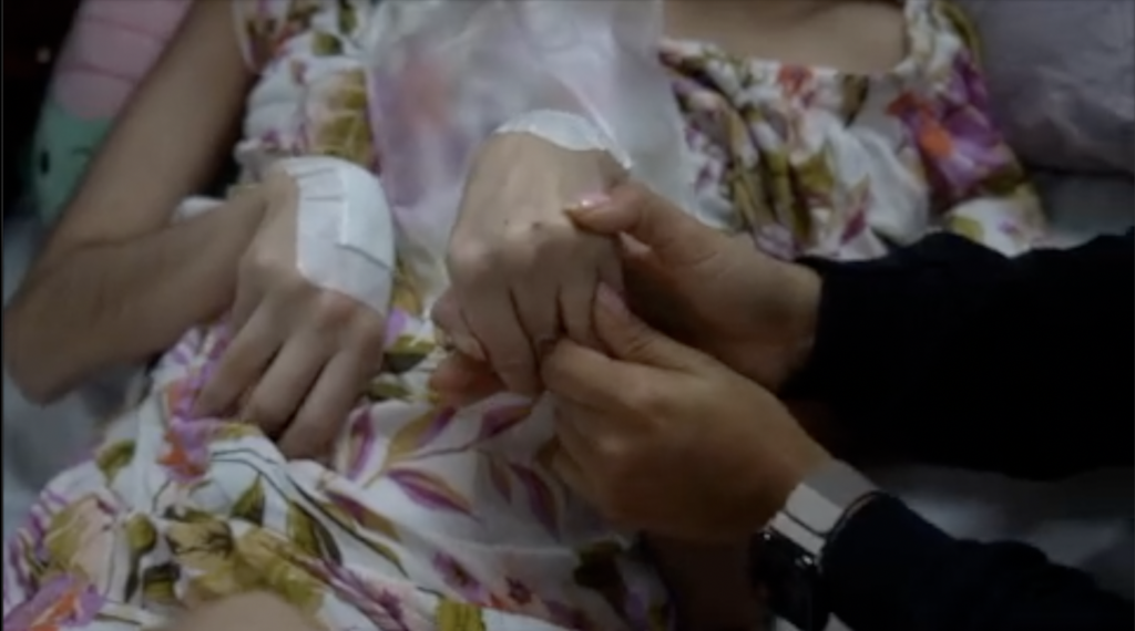 A close up of two pairs of hands against a floral nightie. The mother holds her dying daughter's hands